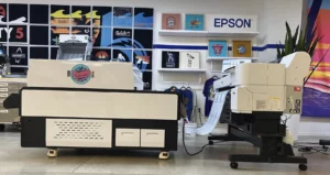Impressora DTF Epson SC-G6070 imprimindo filme que vai direto para o forno batedor de pó (shaker), em sala que imita um ambiente de produção de personalizados.